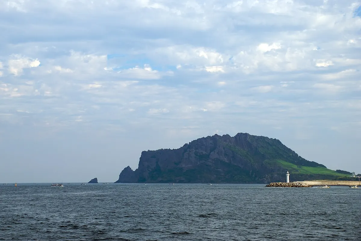 제주 성산일출봉이 바다 위에 솟아 있고 오른쪽 아래에 방파제와 흰 등대가 보이는 풍경